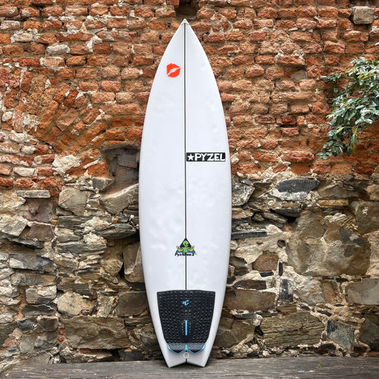 A white Pyzel Pyzalien 2 XL Surfboard with black and blue graphics, standing vertically against a rustic brick and stone wall.