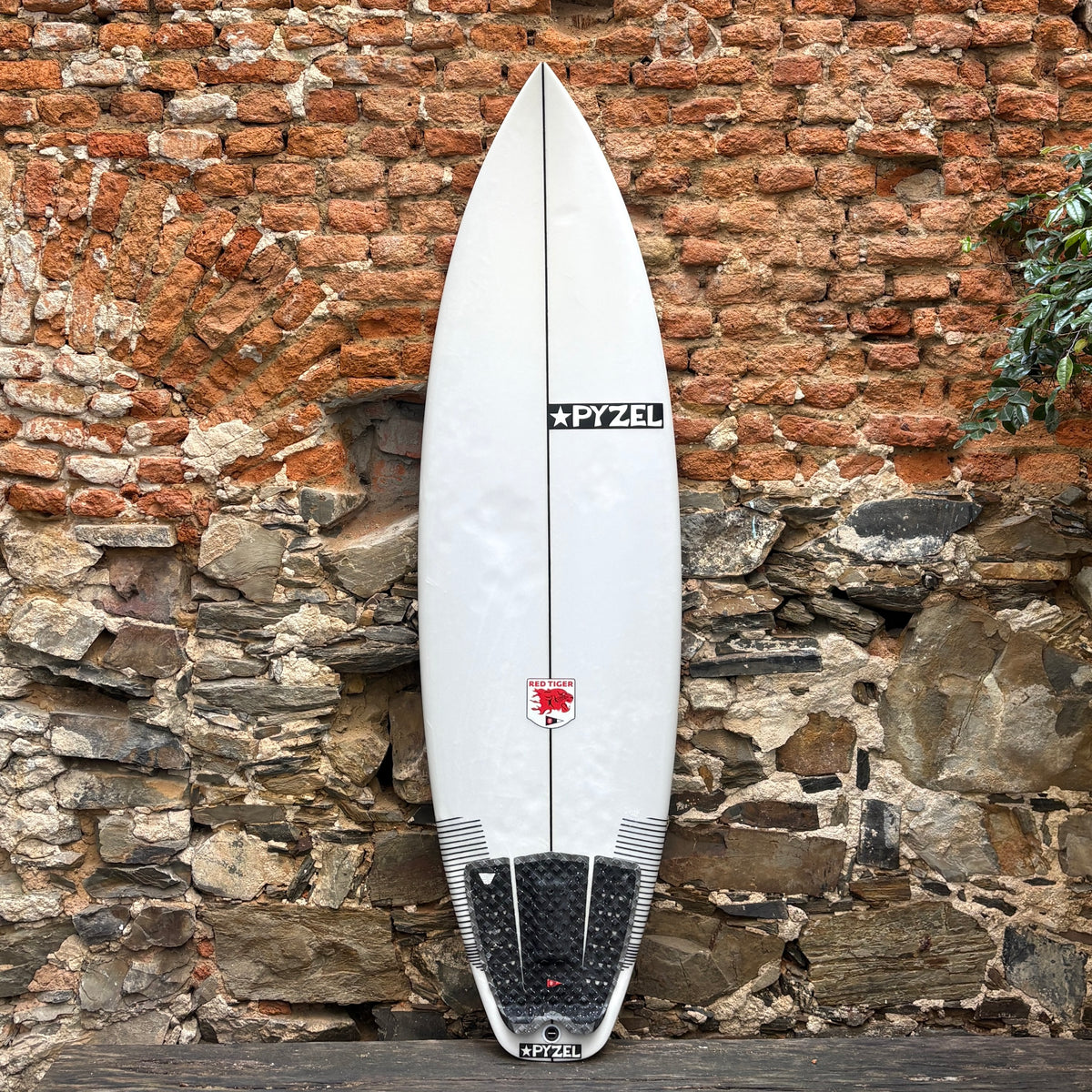 A white surfboard with black and red graphics, including the brand name 'PYZEL' and a red bull logo, leaning against a rustic brick and stone wall.