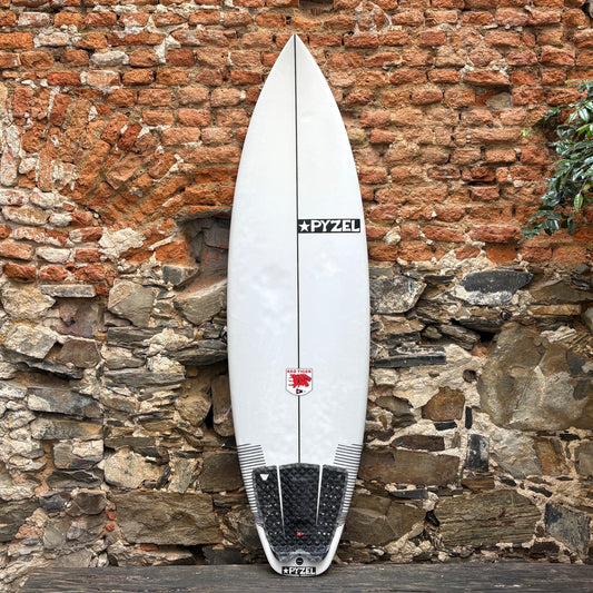 A white surfboard with black and red graphics, including the brand name 'PYZEL' and a red bull logo, leaning against a rustic brick and stone wall.