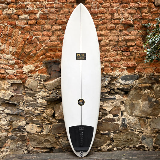 A white surfboard with a black tail pad and a central black line, leaning against a rustic brick and stone wall. The board features a 'PPEEL' brand label and a circular logo near the center.