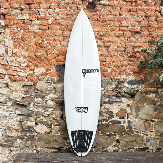 White surfboard with PYZEL SHADOW branding against a rustic stone wall