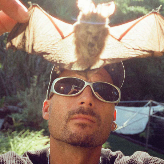 Person wearing sunglasses with a bat perched on their head outdoors