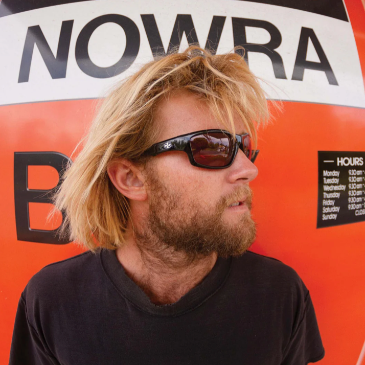 Man with sunglasses and a beard standing in front of a NOWRA sign.