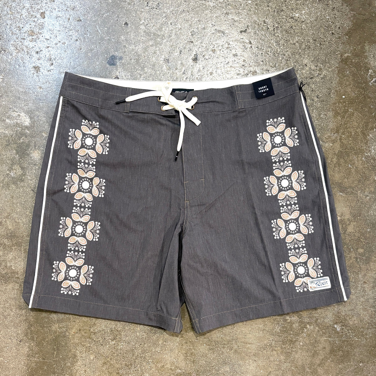 A pair of dark gray board shorts with white and beige geometric patterns down the sides, laid flat on a concrete floor.