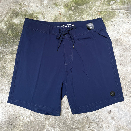 A pair of navy blue RVCA board shorts laid flat on a textured concrete surface. The shorts feature a drawstring waistband with the 'RVCA' brand name printed in white, a side pocket, and a small circular logo patch near the lower left hem.