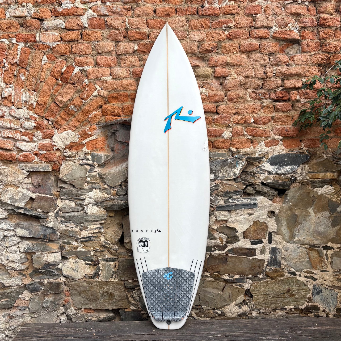 White Rusty surfboard with a blue logo against a stone wall