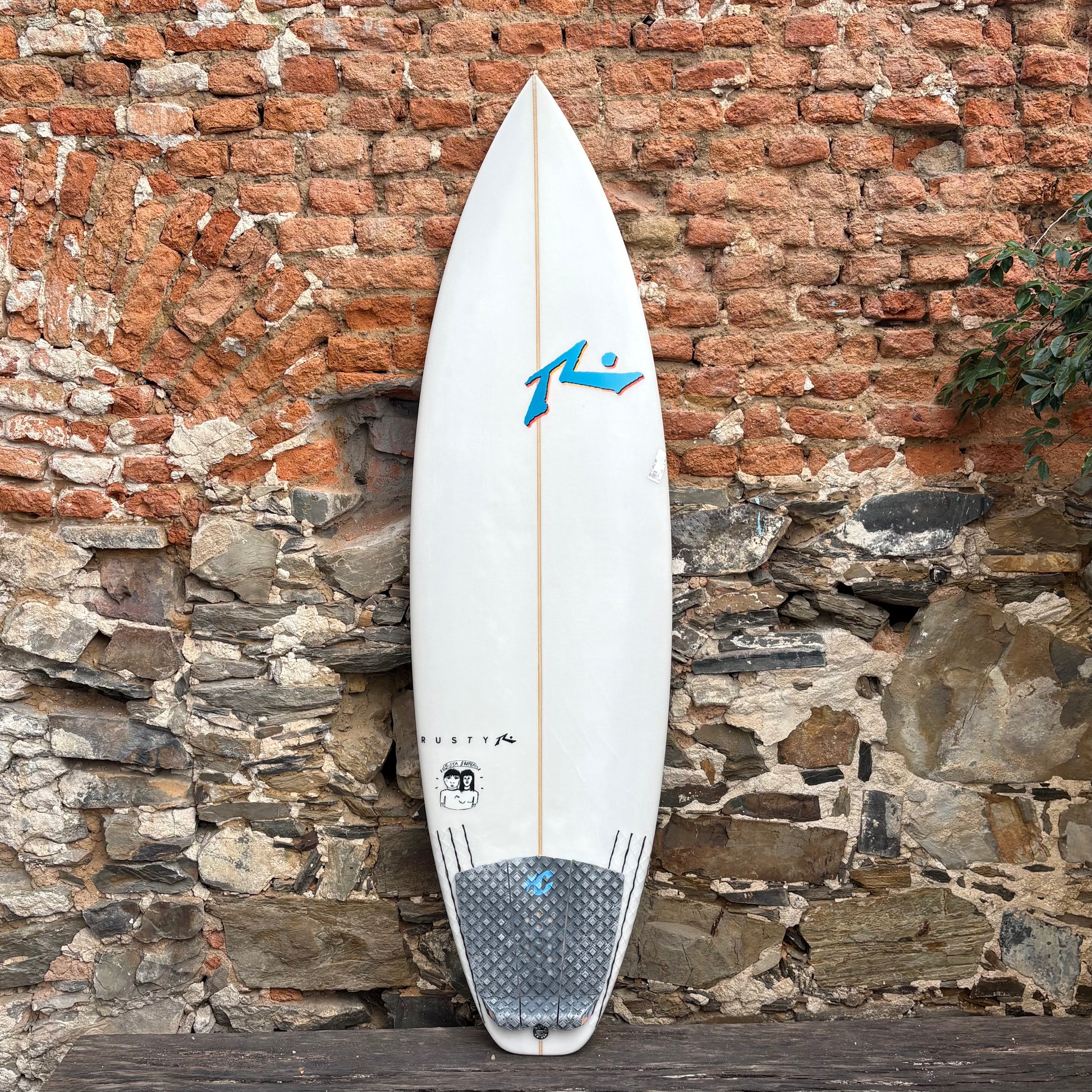 White Rusty surfboard with a blue logo against a stone wall