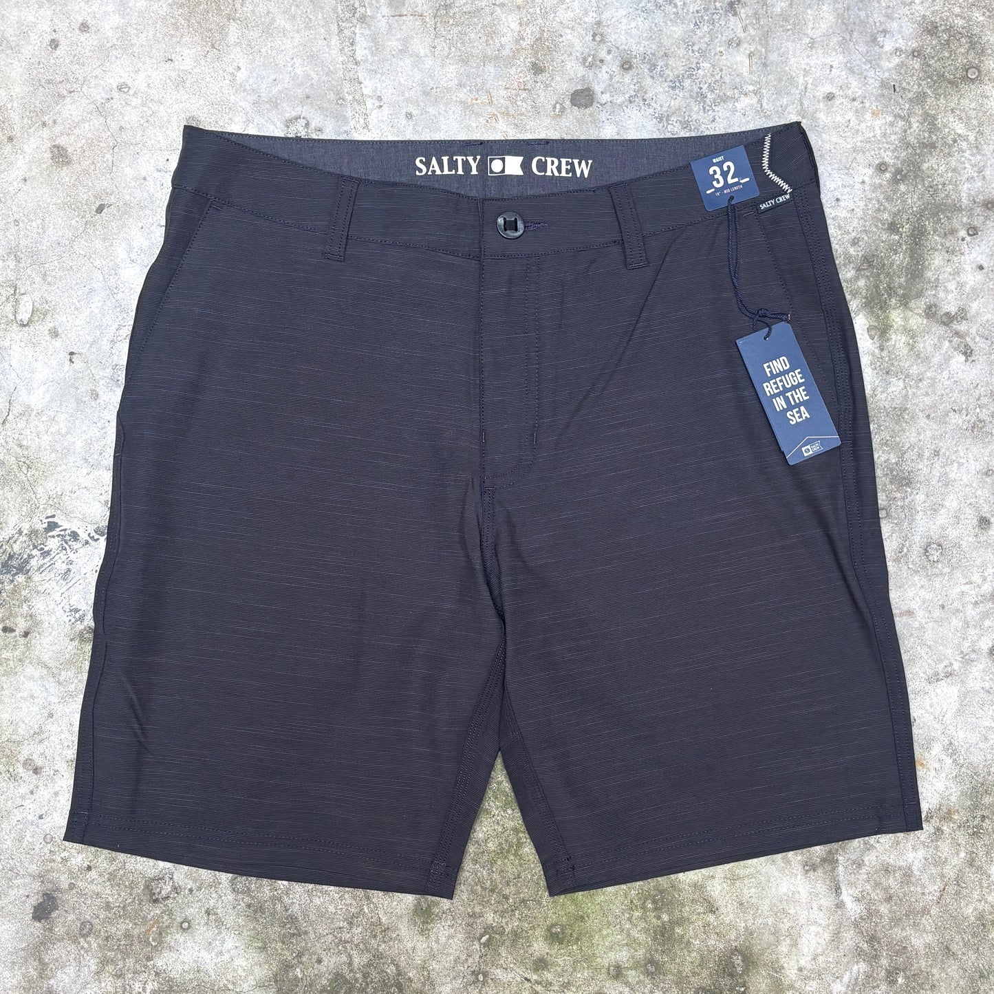 A pair of dark charcoal gray heathered shorts with 'SALTY CREW' printed on the waistband, laid flat on a textured concrete surface with a blue size and care tag attached.