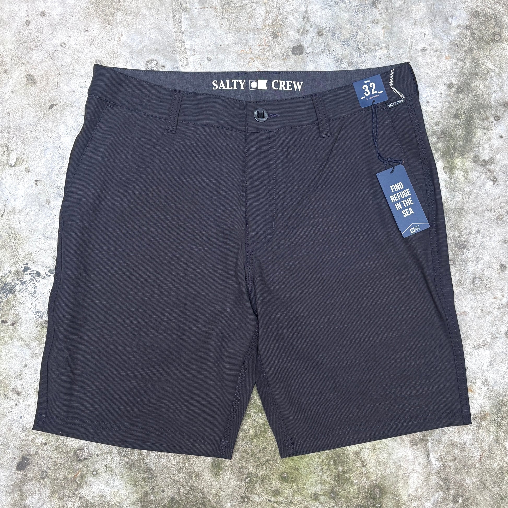 A pair of dark charcoal gray heathered shorts with 'SALTY CREW' printed on the waistband, laid flat on a textured concrete surface with a blue size and care tag attached.