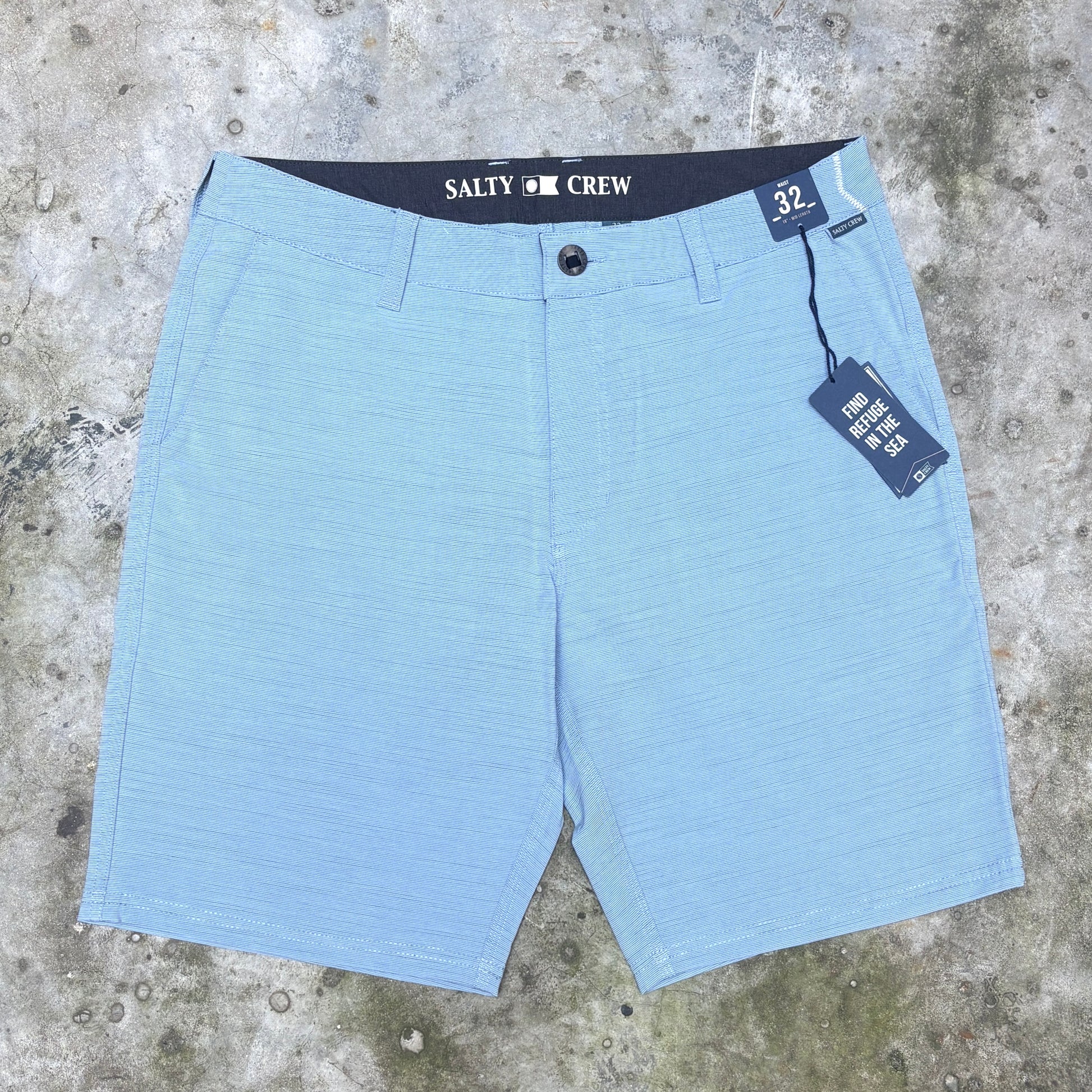 Light blue textured chino shorts with a dark navy waistband featuring the white printed brand name 'SALTY CREW', laid flat on a gray concrete surface with retail tags attached.