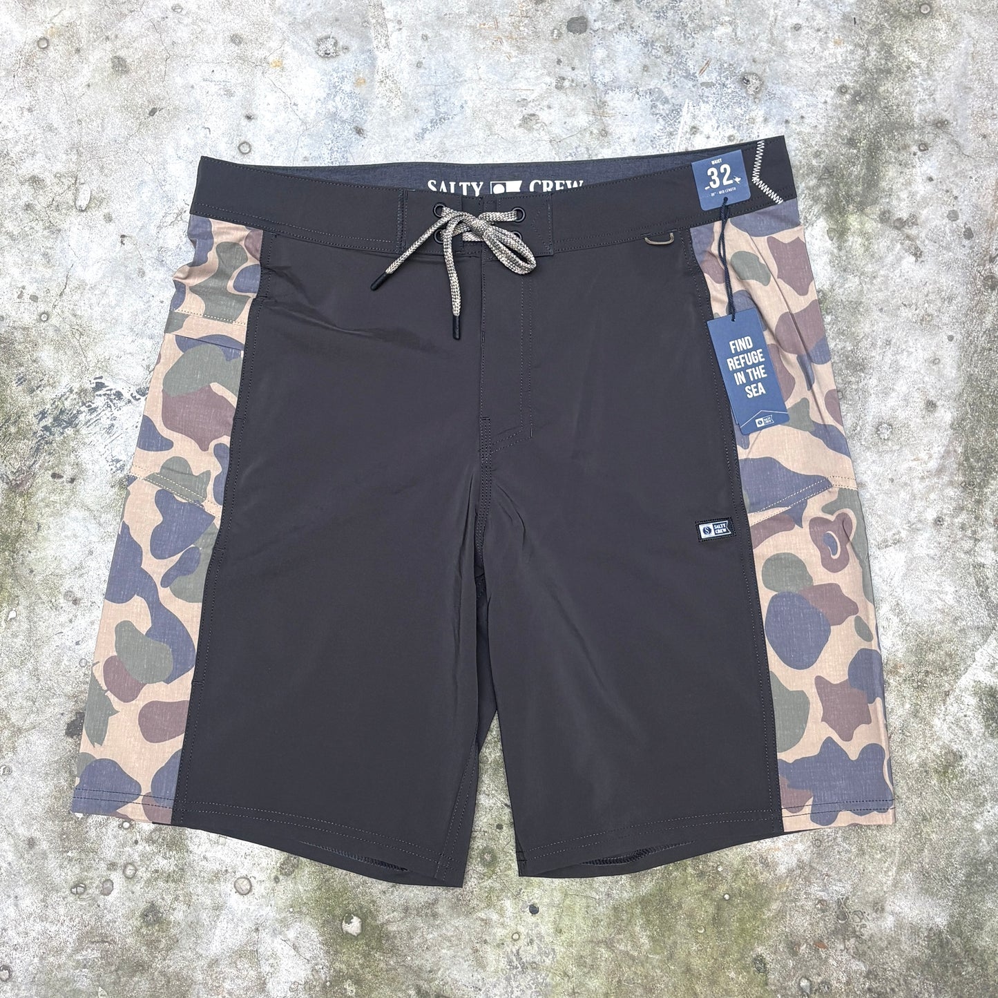 A pair of Salty Crew board shorts laid flat on a textured concrete surface. The shorts feature a solid black front panel, gray drawstring waist, and side panels with a beige, green, and blue camouflage pattern.
