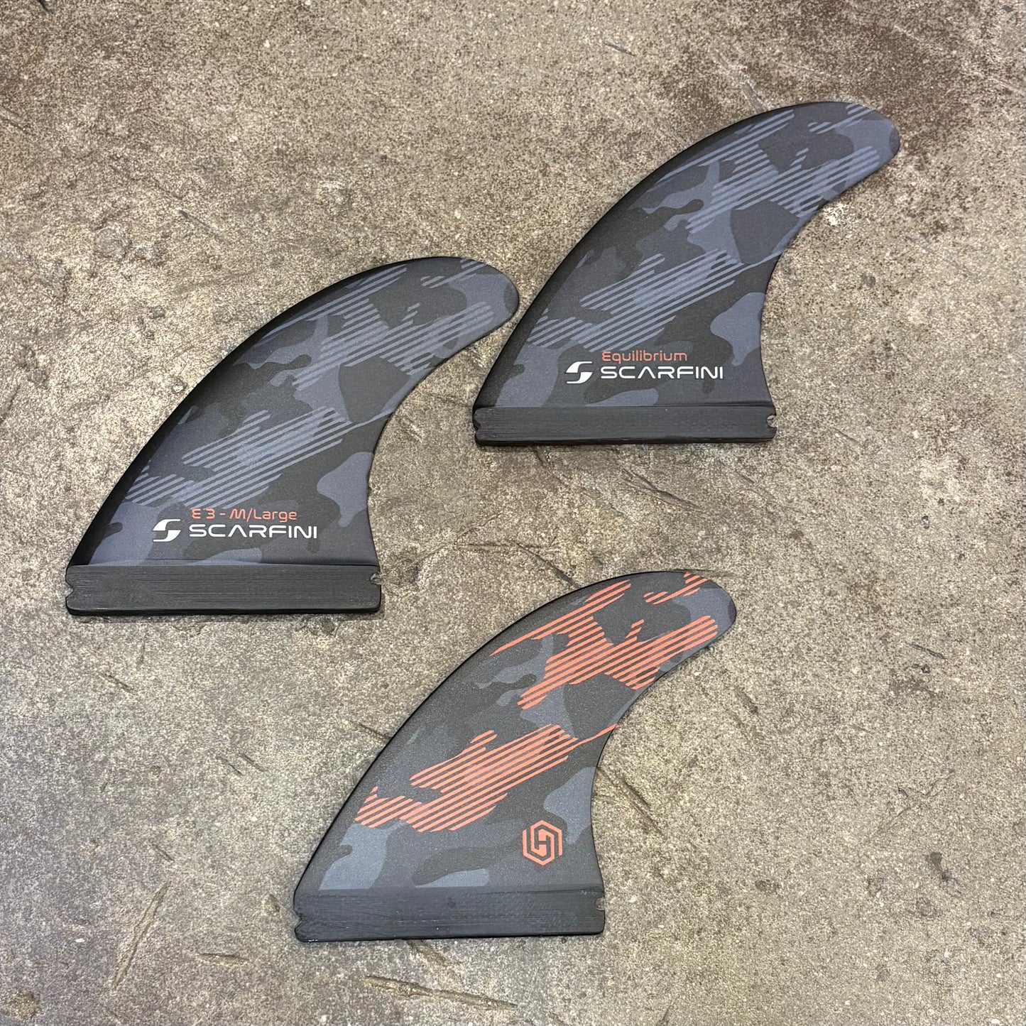 Three surfboard fins with 'Scarfini' branding on a concrete surface
