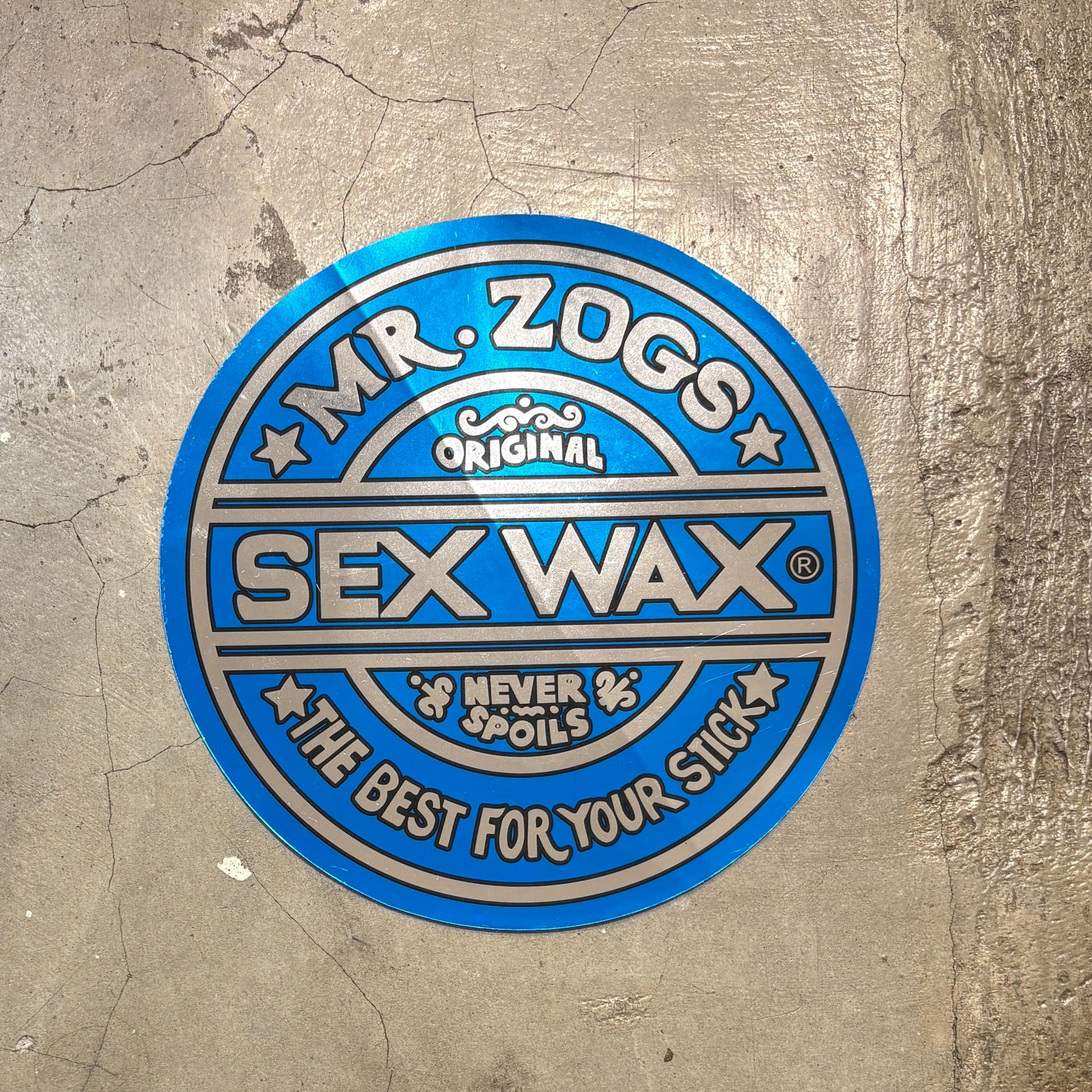 A circular blue and silver sticker for 'Mr. Zogs Sex Wax' is shown on a textured concrete floor.