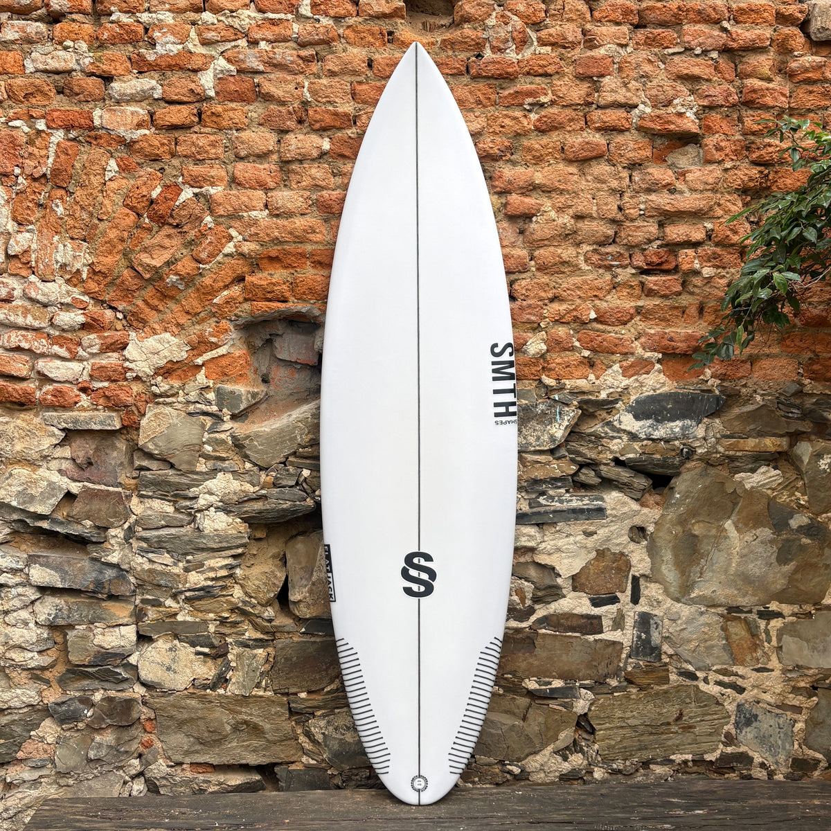A white surfboard with black graphics and the brand name 'SMITH' printed vertically, leaning against a rustic brick and stone wall.