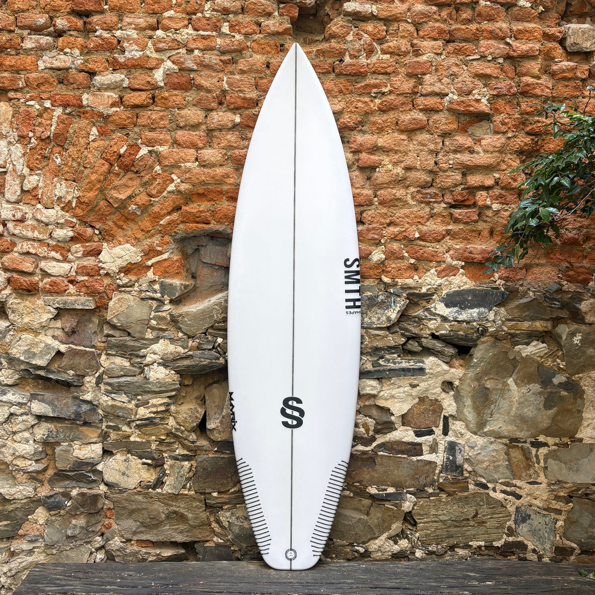 A white surfboard with black graphics and the brand name 'SMITH' printed vertically, leaning against a rustic brick and stone wall.