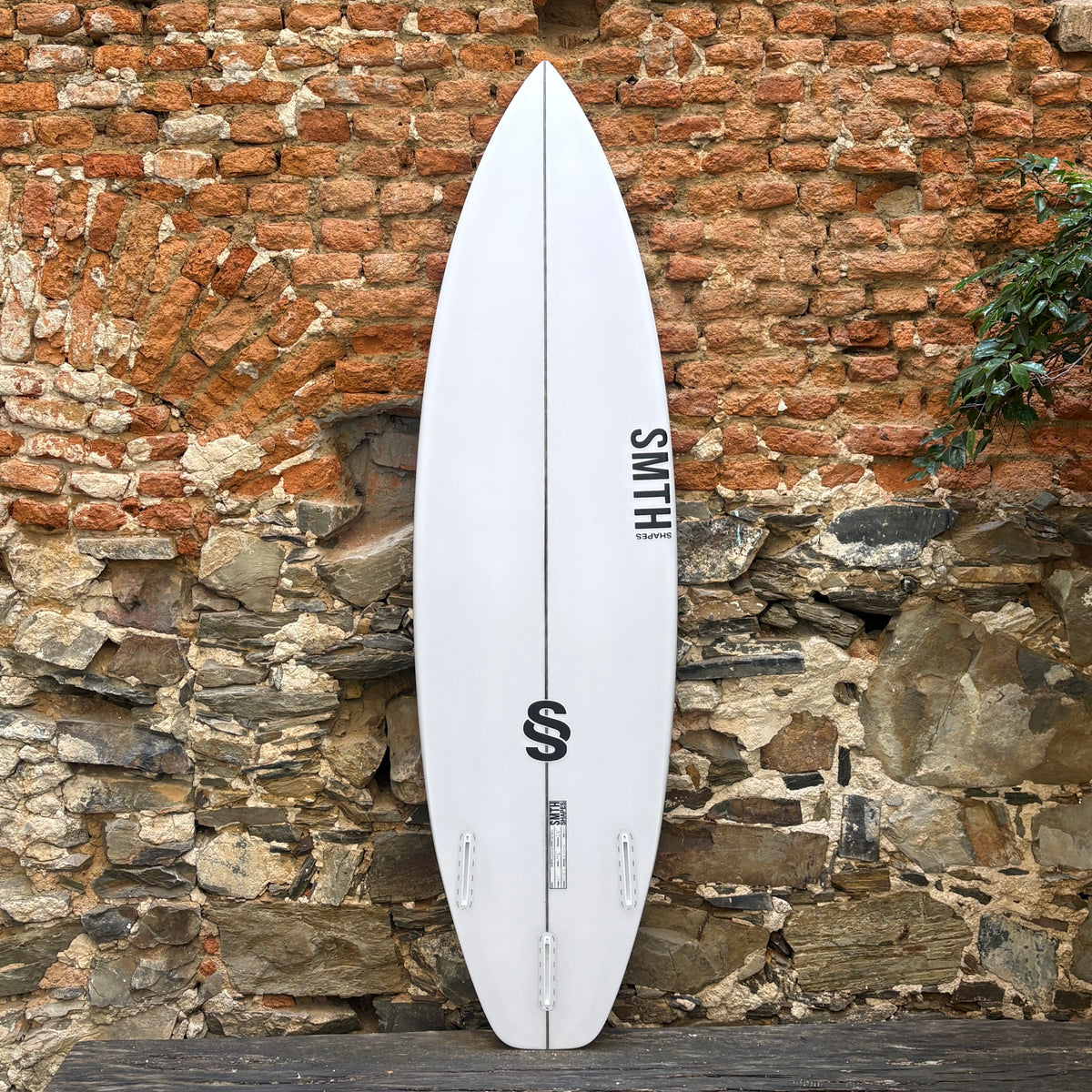 A white surfboard with black graphics and the brand name 'SMITH' printed vertically, leaning against a rustic brick and stone wall.