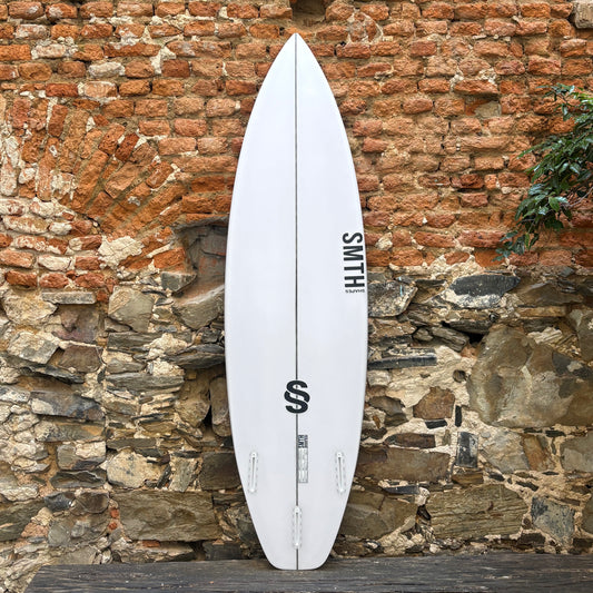 A white surfboard with black graphics and the brand name 'SMITH' printed vertically, leaning against a rustic brick and stone wall.