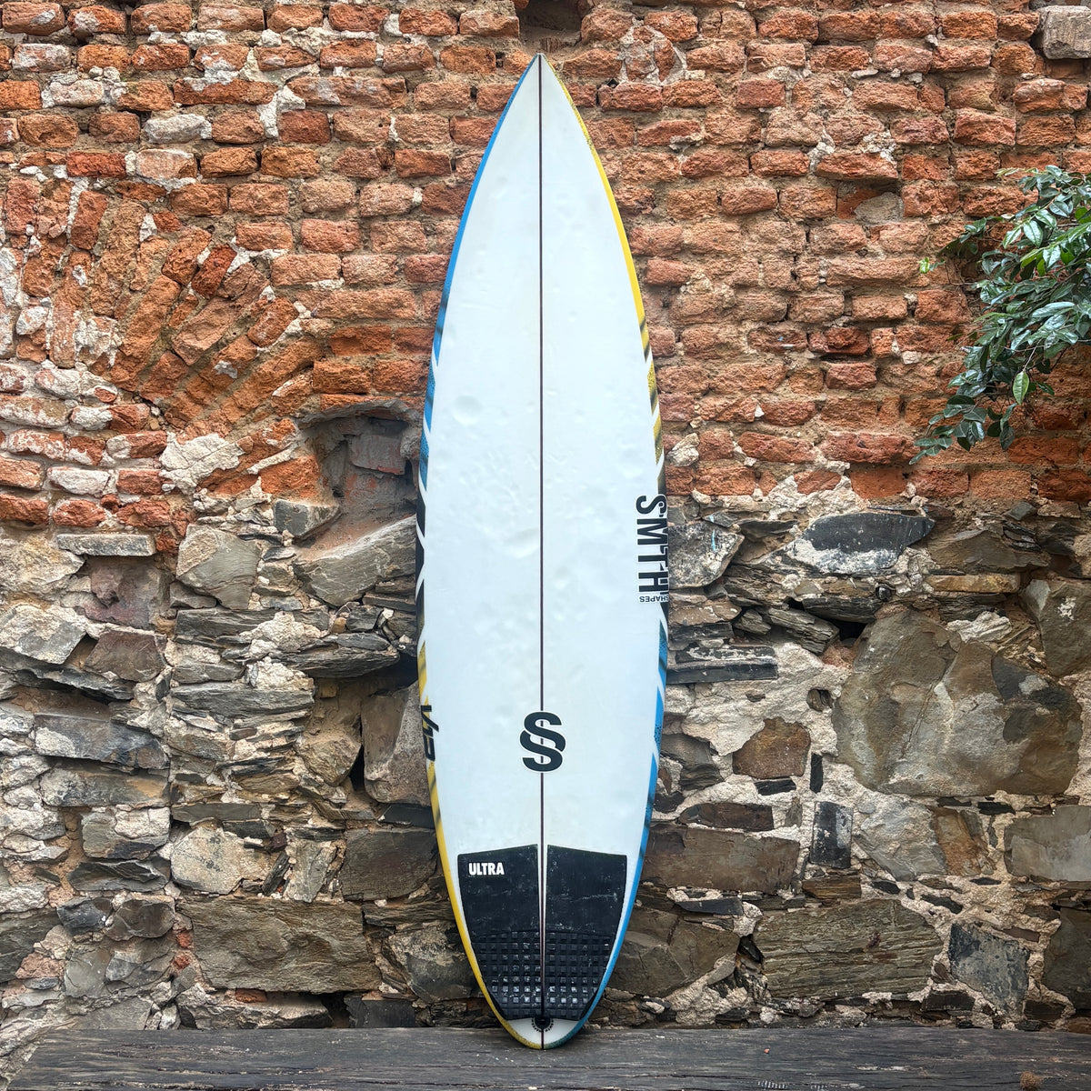A surfboard with a white base, blue and yellow edge stripes, and black graphics, leaning against a rustic brick and stone wall.