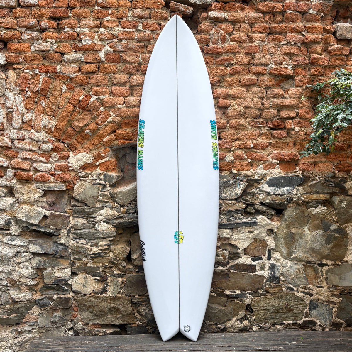 A white surfboard with blue and yellow graphics, leaning against a rustic brick and stone wall.