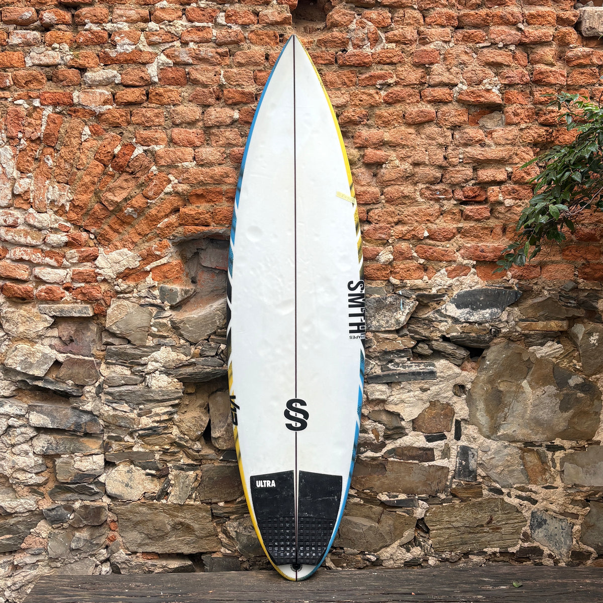 A surfboard with a white deck, yellow and blue edge stripes, and a black tail pad, leaning against a rustic brick and stone wall.