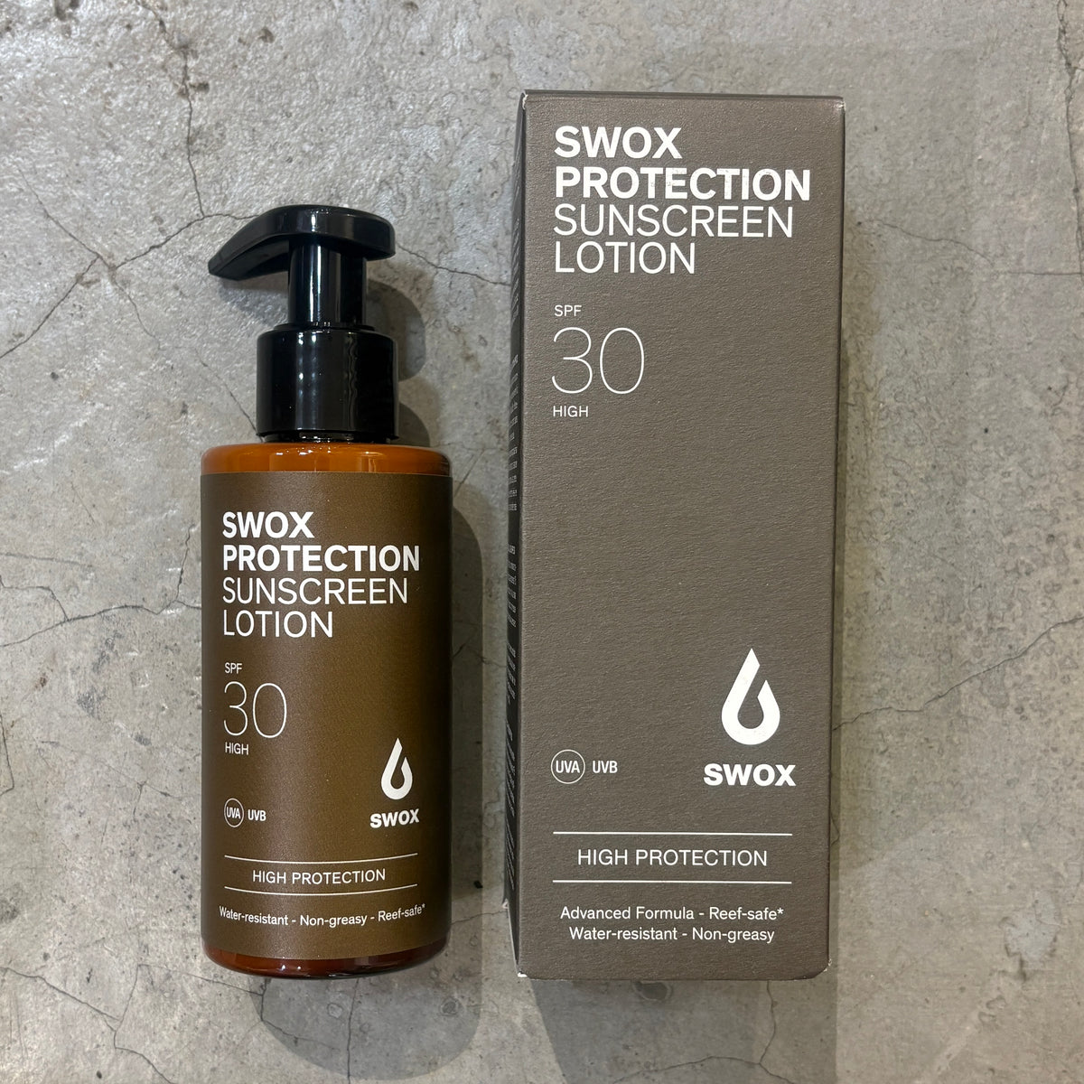 A bottle of Swox Protection Sunscreen Lotion SPF 30 next to its matching cardboard box, displayed on a textured gray stone surface.