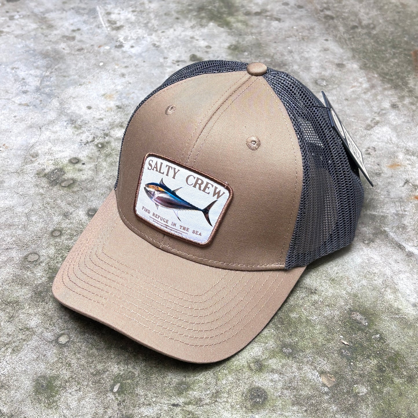 A tan and grey trucker hat with a 'Salty Crew' fish patch on the front, resting on a textured concrete surface.