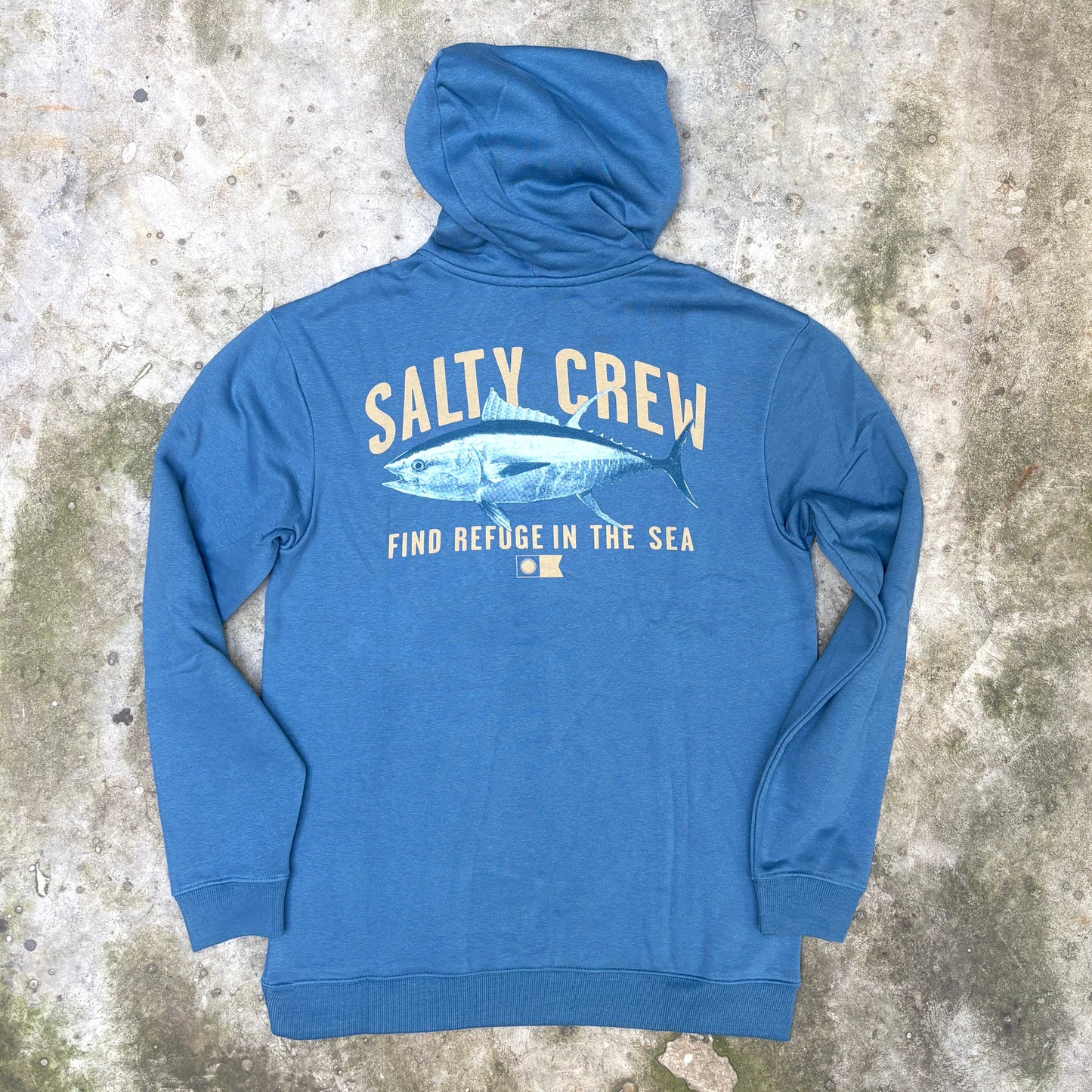 A blue hoodie laid flat on a textured concrete surface, showing the back side with a large graphic print that reads 'SALTY CREW' and 'FIND REFUGE IN THE SEA' with a tuna fish illustration.