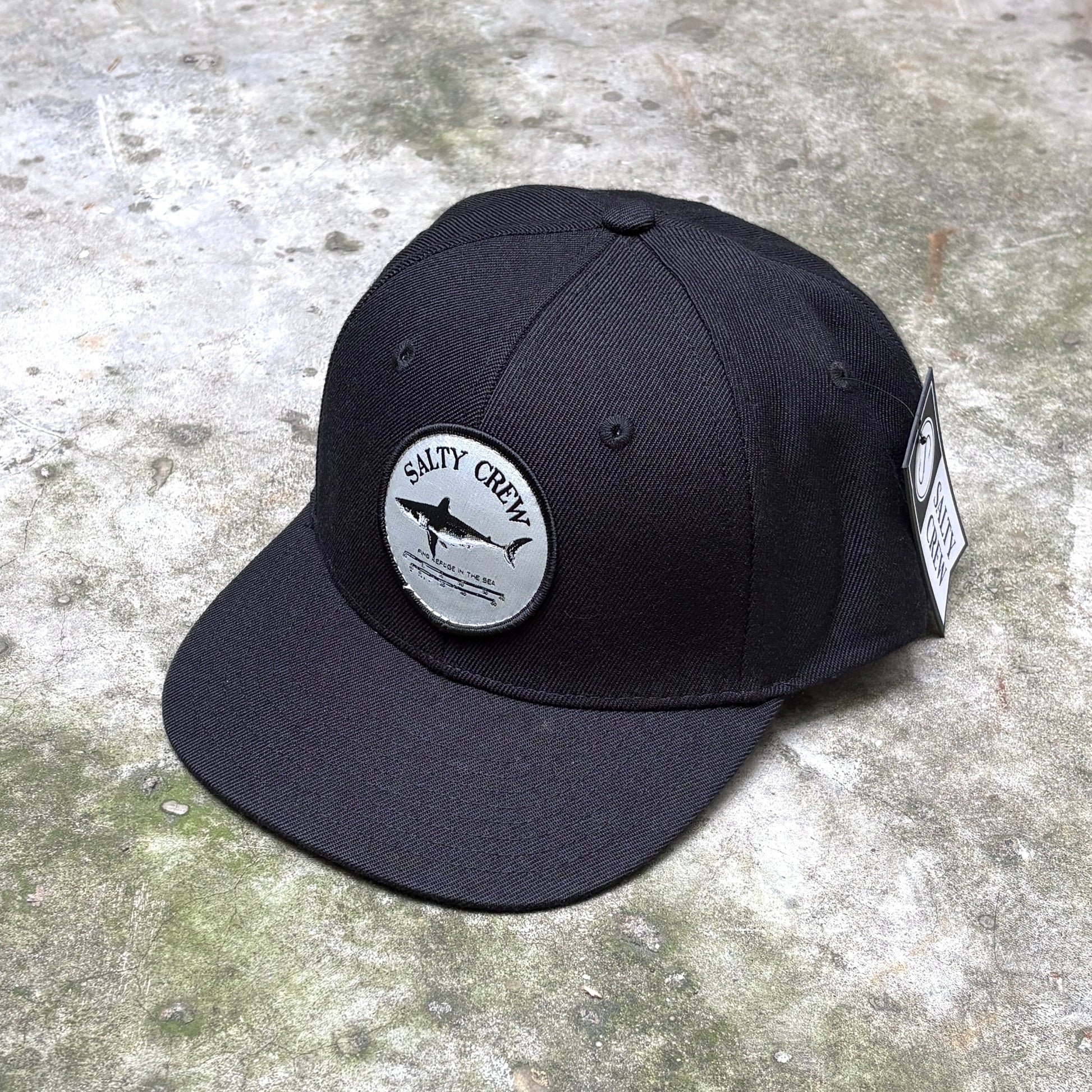 A black flat-brim baseball cap with a circular gray patch on the front that reads 'Salty Crew' with a shark graphic, photographed on a textured concrete surface.