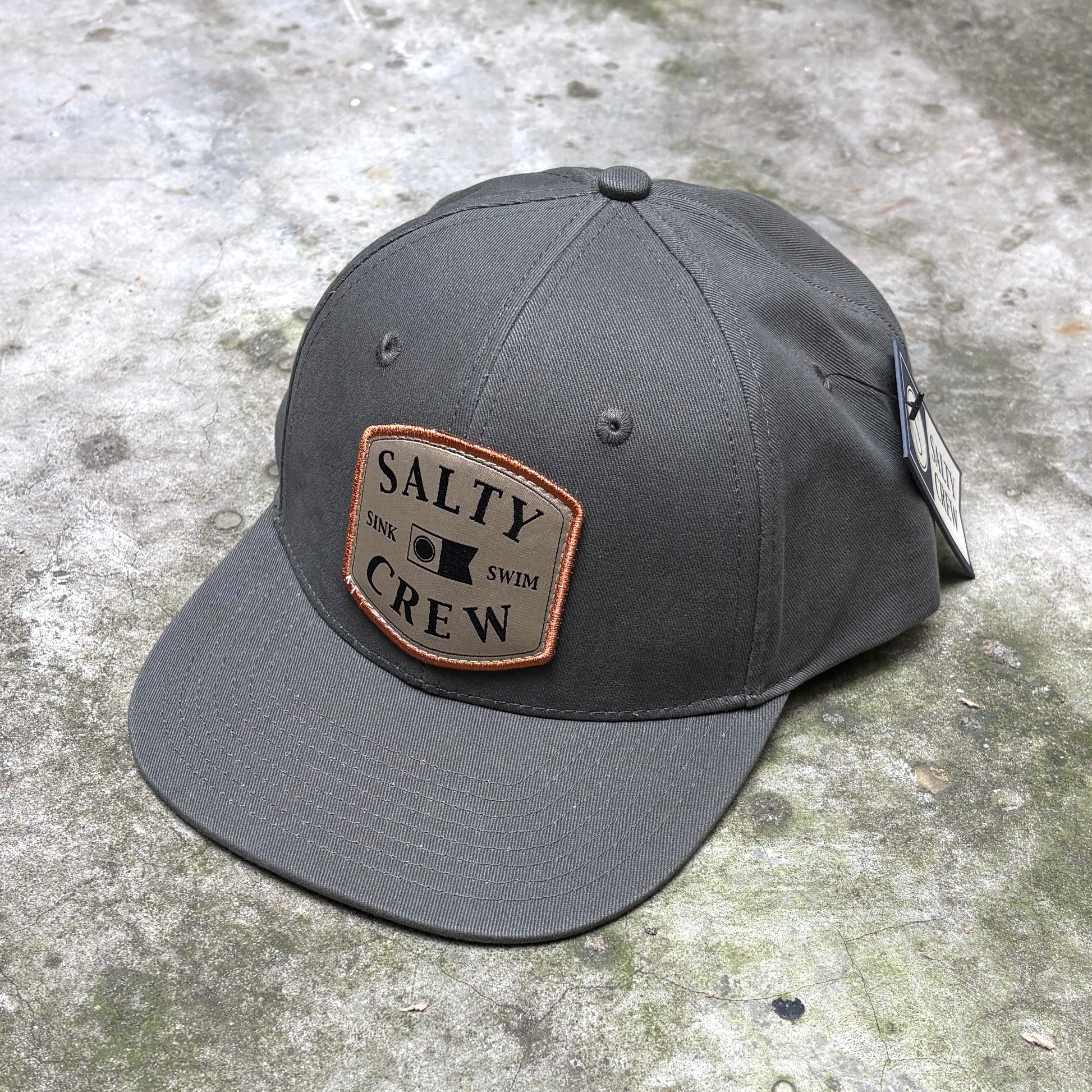 A dark gray snapback cap with a tan rectangular patch on the front that reads 'SALTY DINK SWIM CREW' with a small black square logo, photographed on a textured concrete surface.