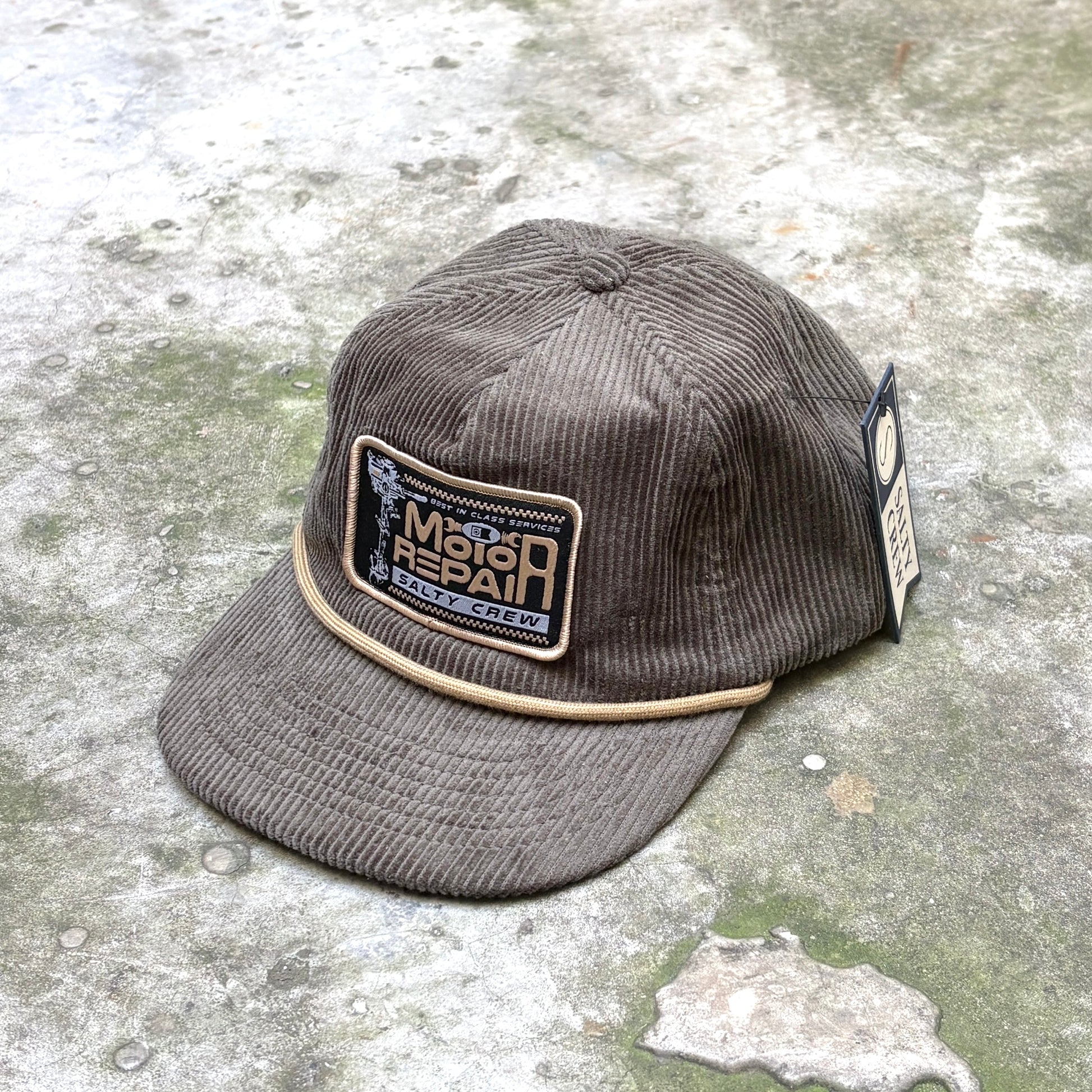A brown corduroy baseball cap with a beige embroidered patch on the front reading 'Mega Repair' and a small 'KAVU' logo, resting on a textured concrete surface.