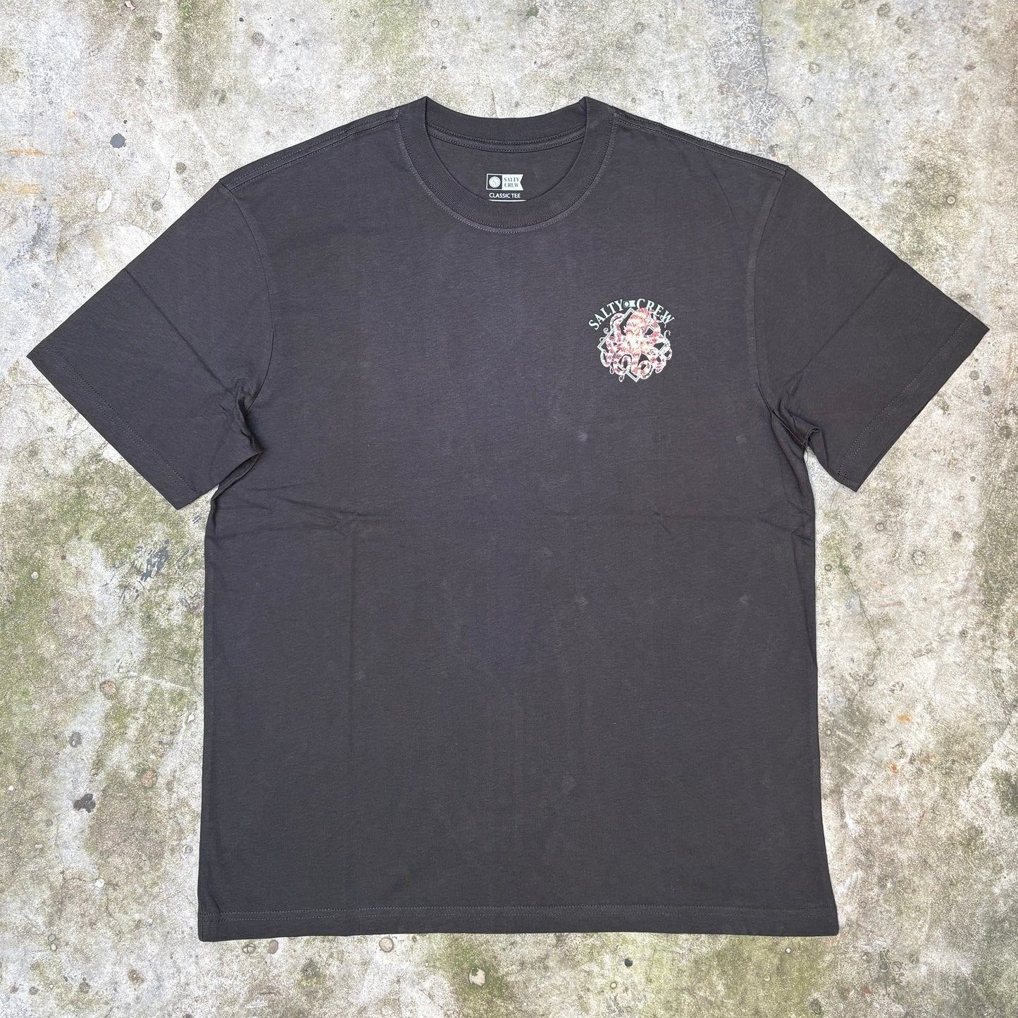 A black short-sleeve t-shirt with a small pink and white graphic on the left chest, laid flat on a textured concrete surface.