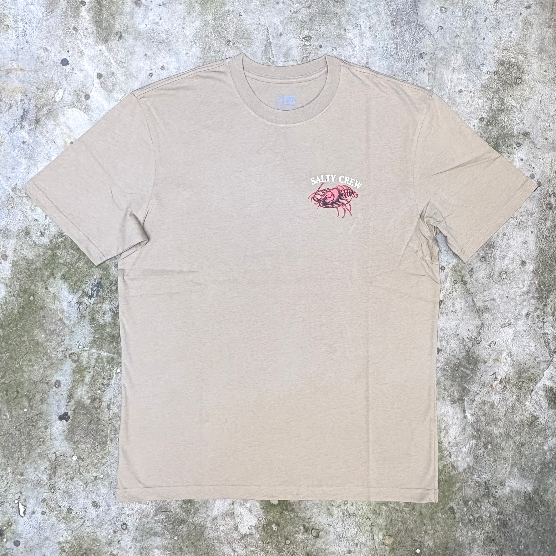 A beige crewneck t-shirt with a small pink and white graphic on the left chest, laid flat on a textured gray concrete surface.