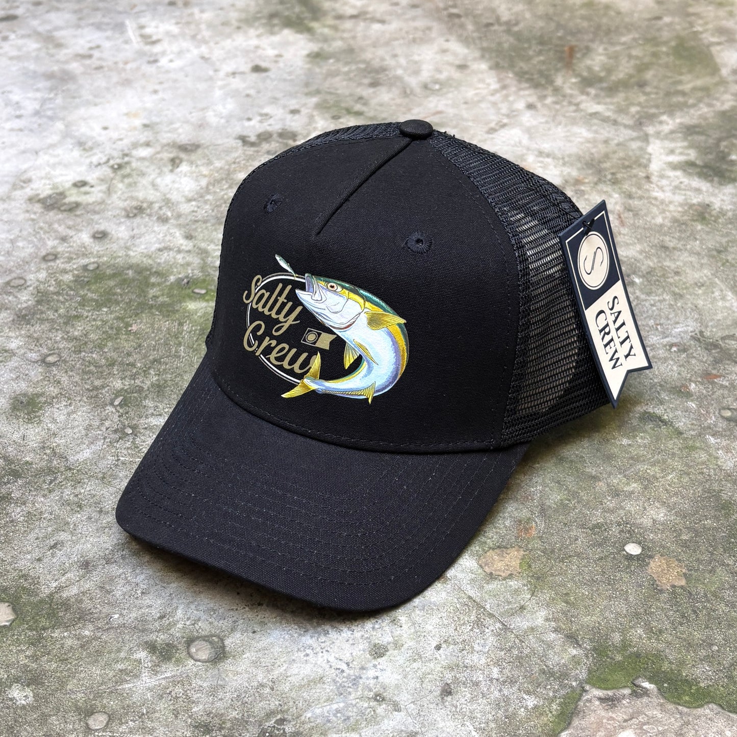 A black trucker hat with a fish graphic and 'Salty Crew' text on the front, resting on a textured concrete surface. A 'Salty Crew' brand tag is attached to the side mesh.
