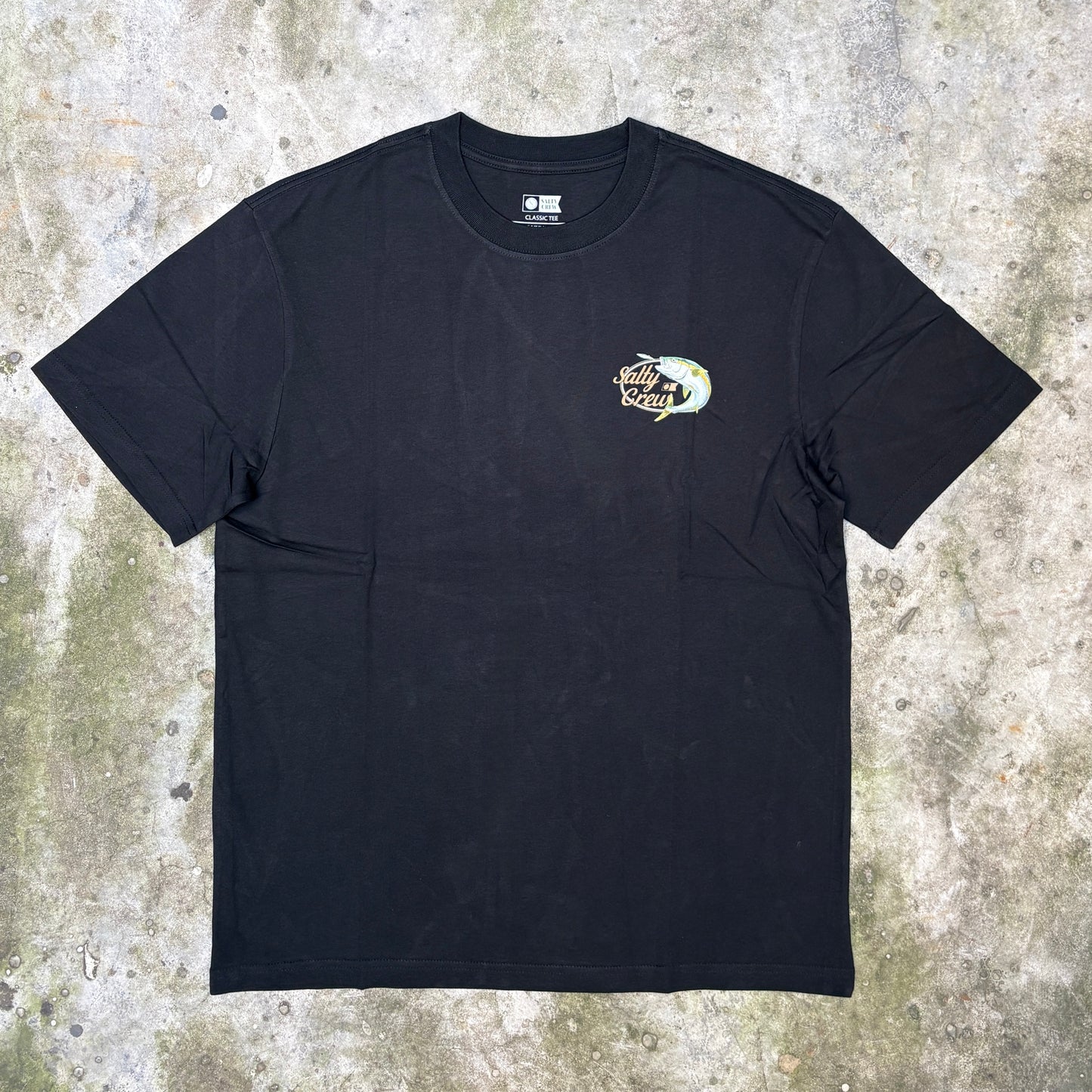 A black short-sleeve crewneck t-shirt with a small multicolor graphic on the left chest, laid flat on a textured concrete surface.