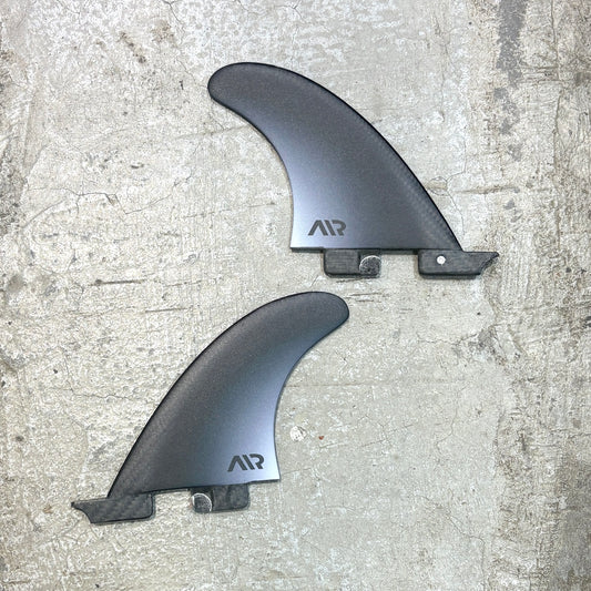 Two dark gray carbon fiber surfboard fins with the AIR logo, displayed on a textured concrete surface.