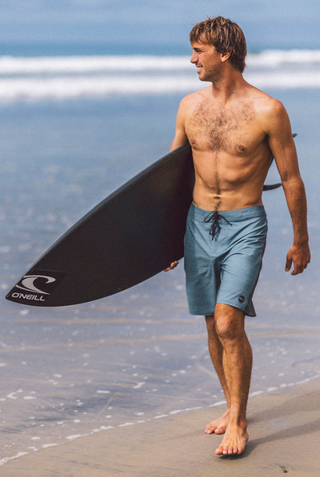 O'Neill Hyperfreak TRVLR boardshorts worn by a shirtless man on the beach carrying a surfboard.