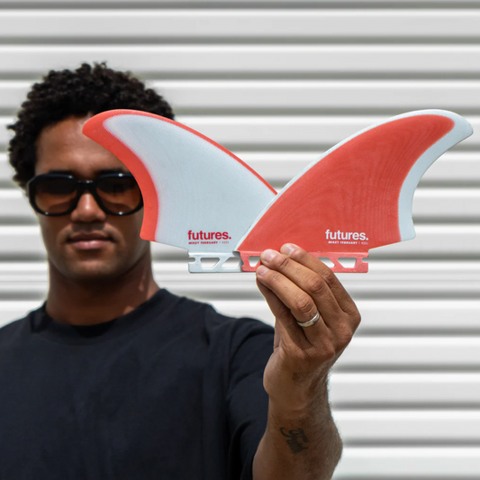 Person holding a red and white surf fin with 'futures' branding against a corrugated metal background.