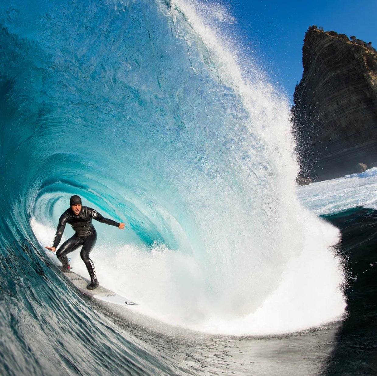 Surfer riding a large wave with a clear blue sky and rocky cliff in the background