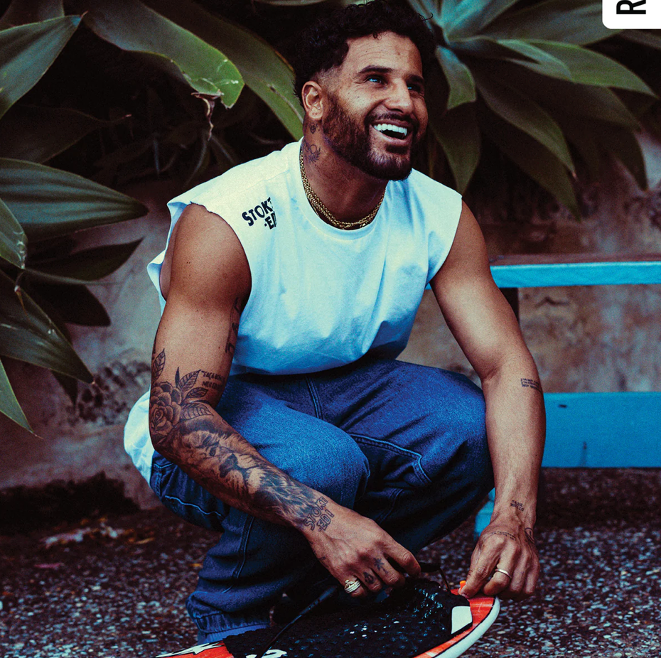 Man sitting on the ground with tattoos, wearing a light blue sleeveless shirt and blue jeans, surrounded by plants.