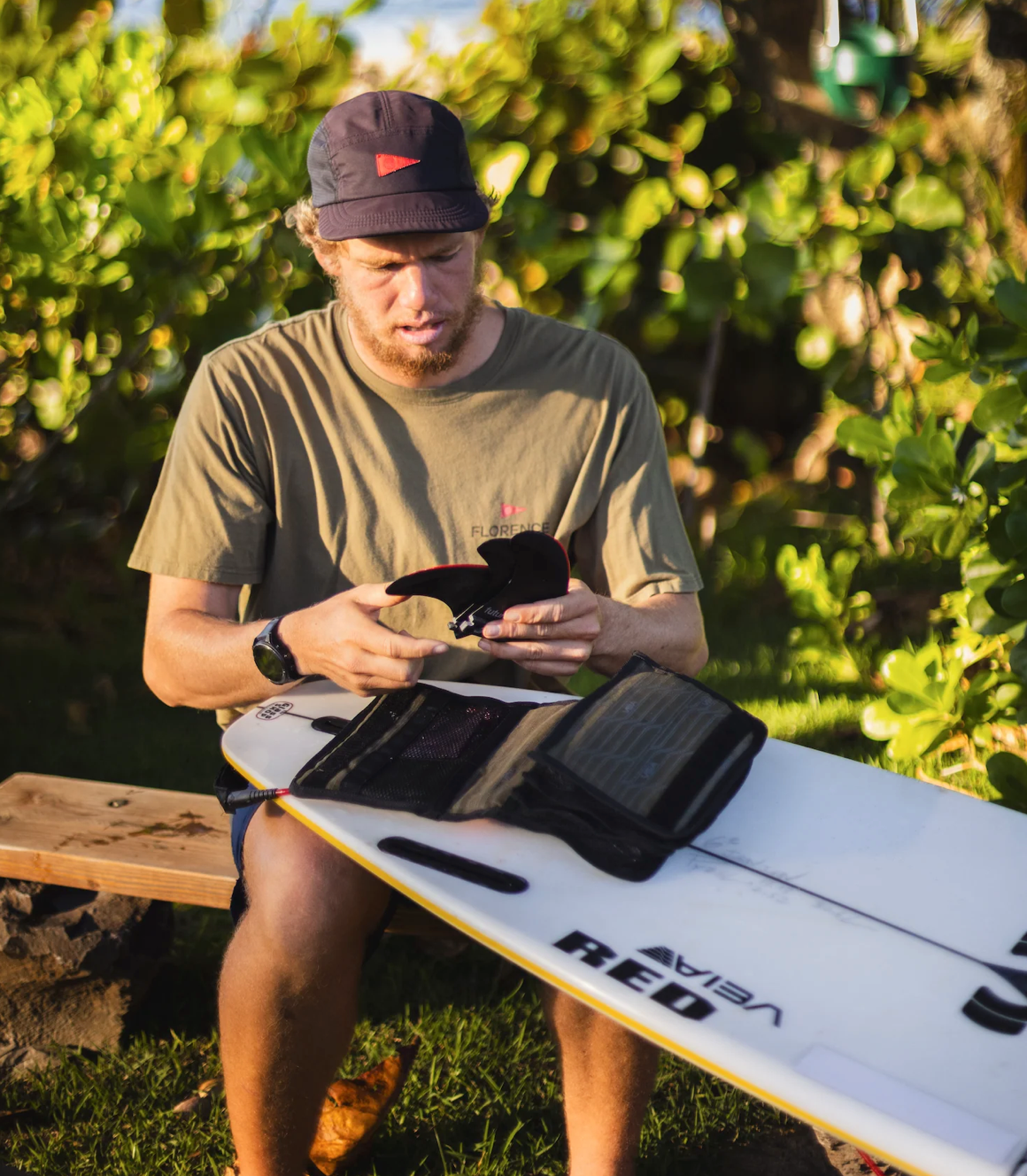 A man sitting outdoors on a wooden bench holding a white surfboard with a black fin and leash attached, wearing an olive green t-shirt and a dark cap.