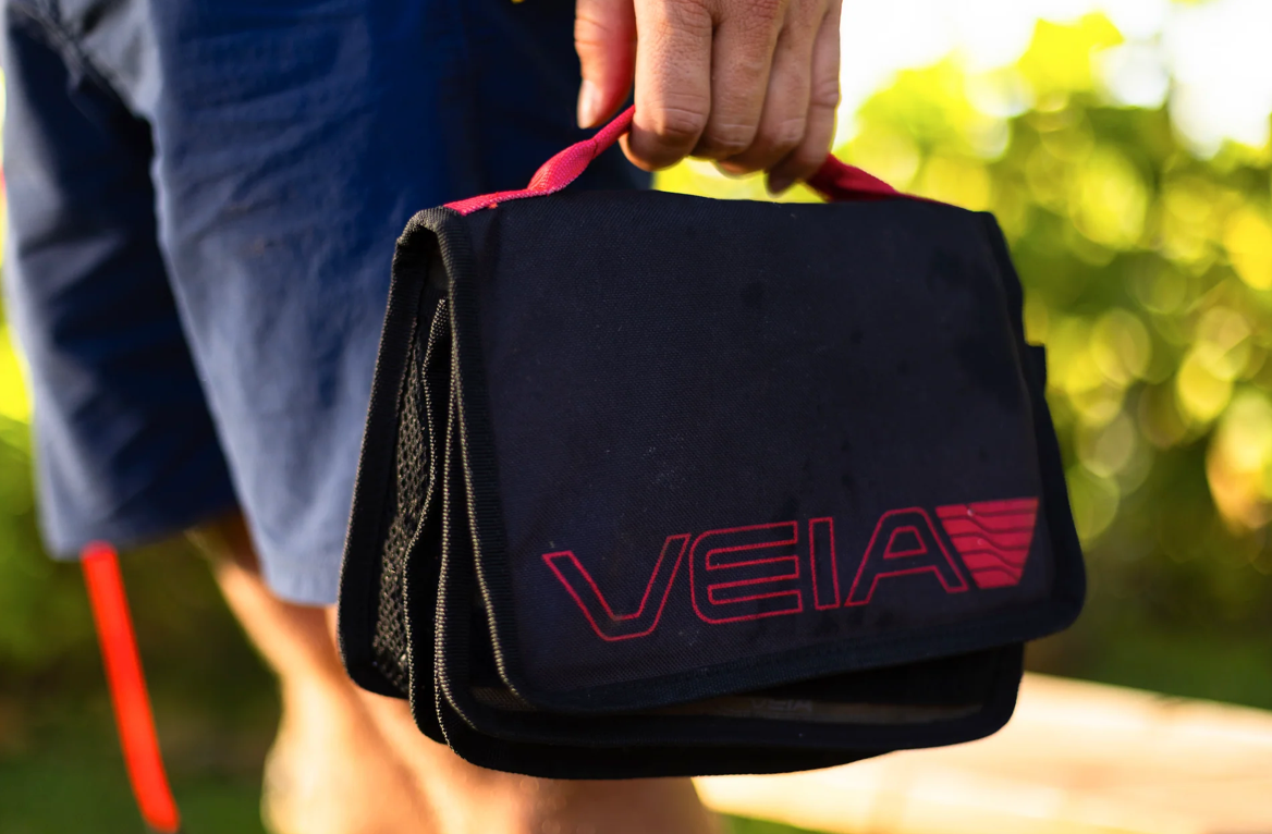 A person outdoors holding a black VEIA storage bag with red trim and a red logo, shown in a close-up shot.