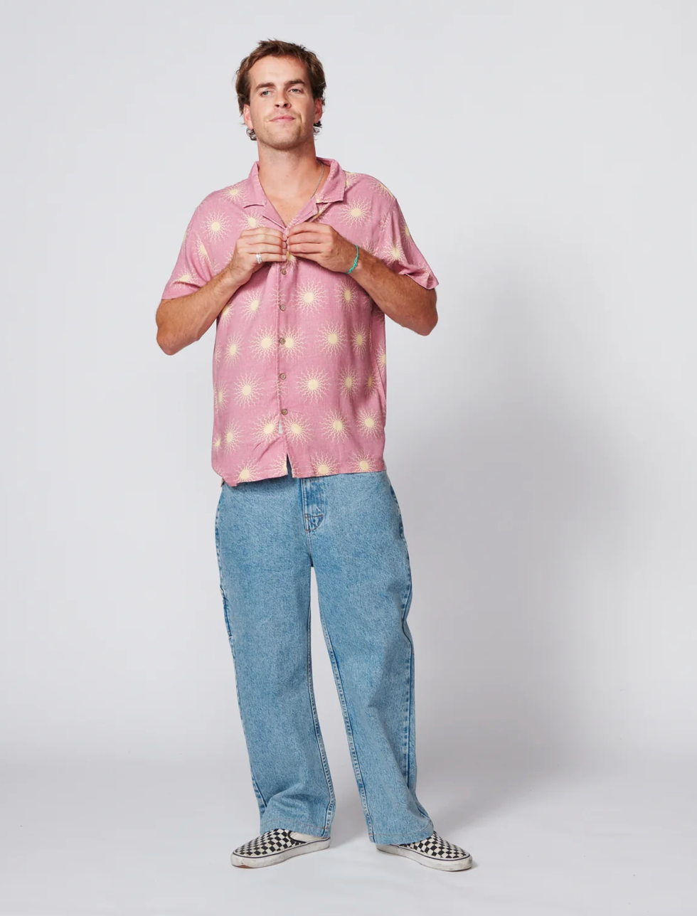 A man modeling a pink short-sleeve button-up shirt with a yellow sunburst pattern, paired with light blue jeans and black-and-white checkered slip-on shoes, against a light gray studio background.