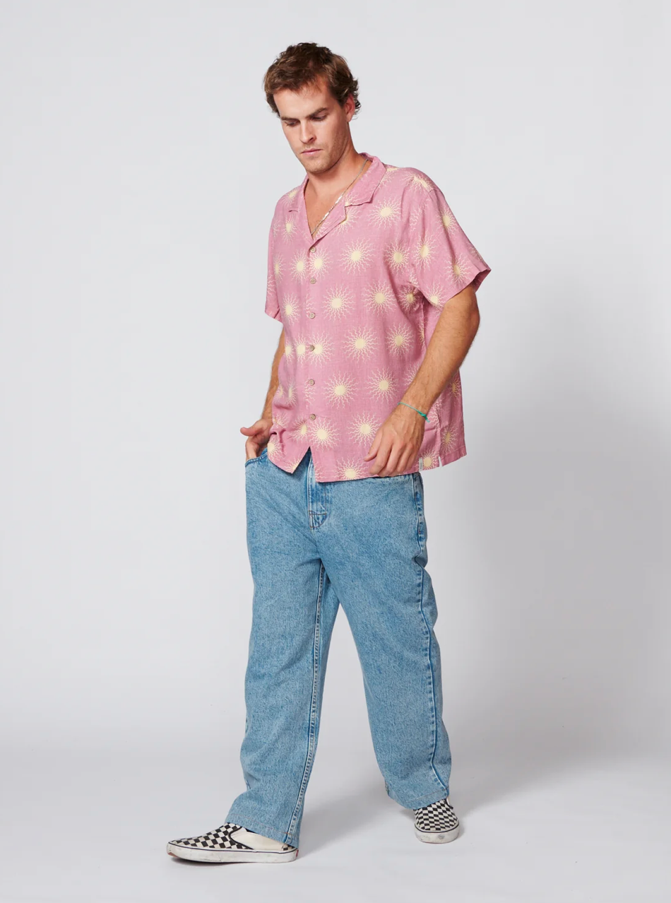 A man modeling a pink short-sleeve button-up shirt with a yellow sunburst pattern, paired with light blue jeans and black-and-white checkered slip-on shoes, against a light gray studio background.