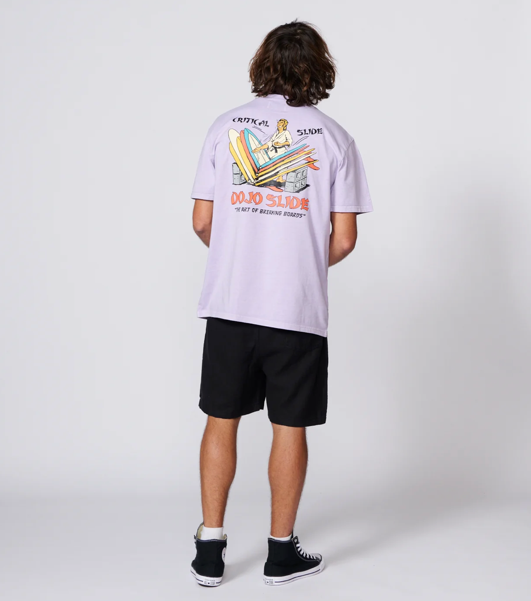 A model shown from the back wearing a light purple short-sleeve polo shirt with a large graphic print on the back, paired with black shorts and black high-top sneakers, against a light gray studio background.
