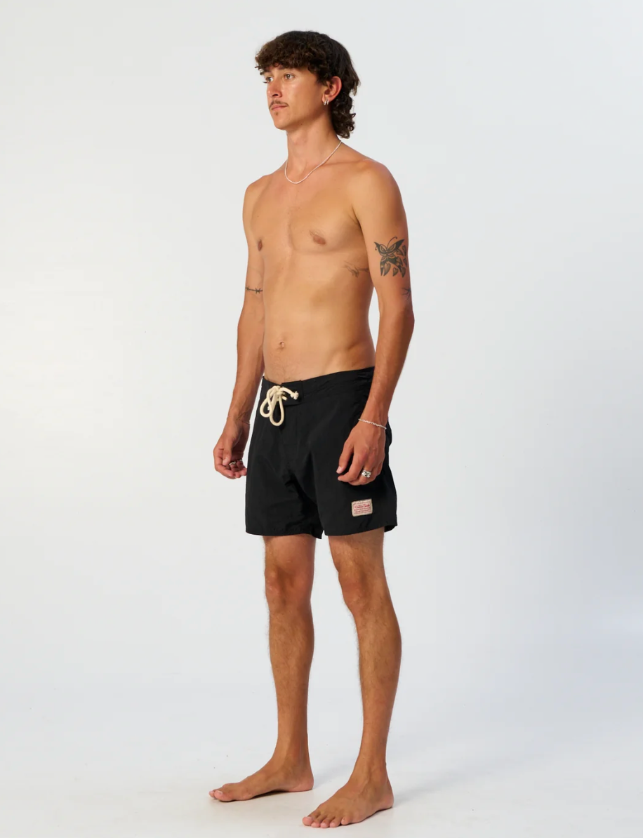 A man modeling black swim trunks with a cream-colored drawstring, standing against a plain white studio background.