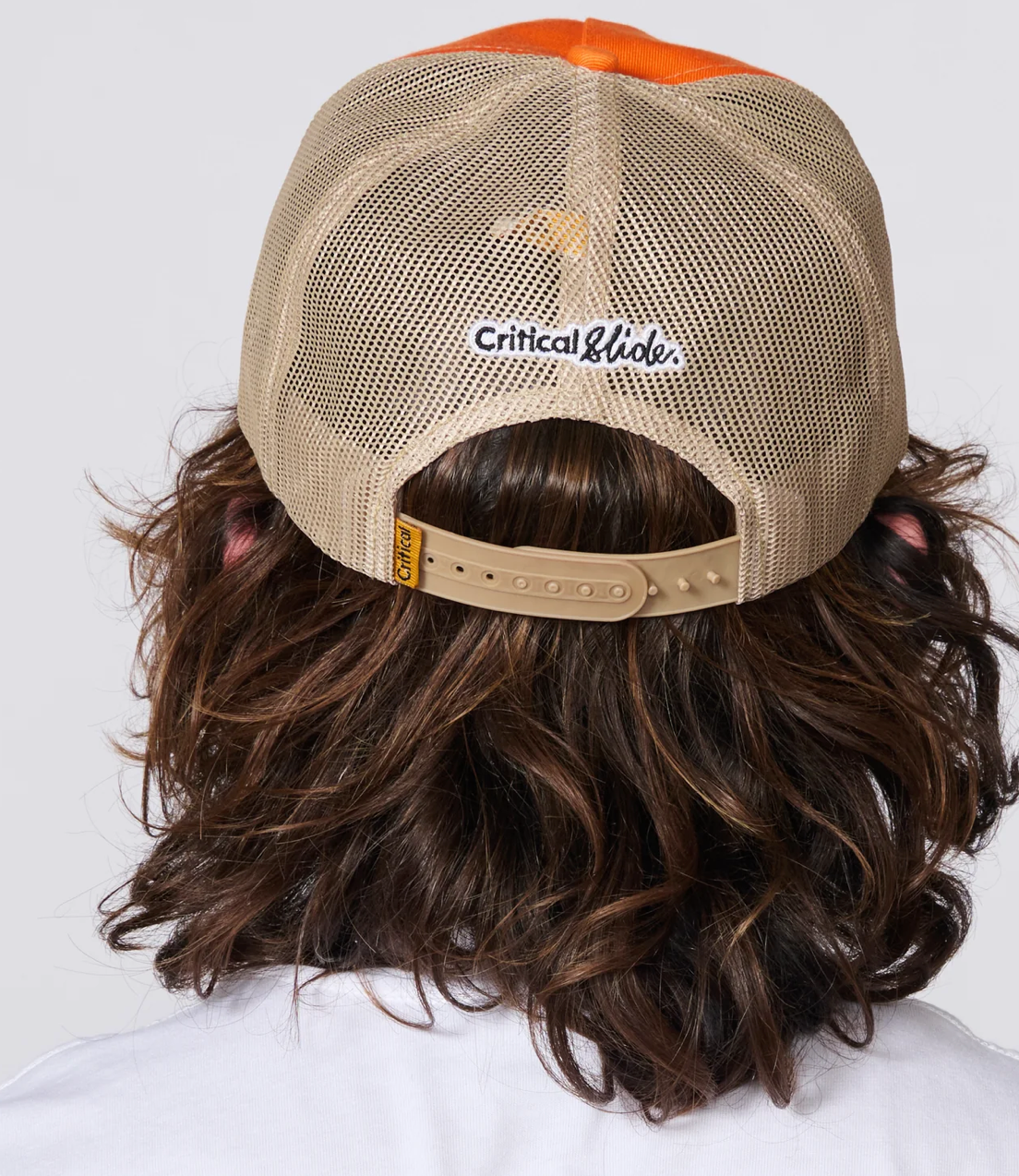 Rear view of a person with wavy brown hair wearing a tan mesh baseball cap with an orange top panel and a white embroidered Critical Glide logo on the back.
