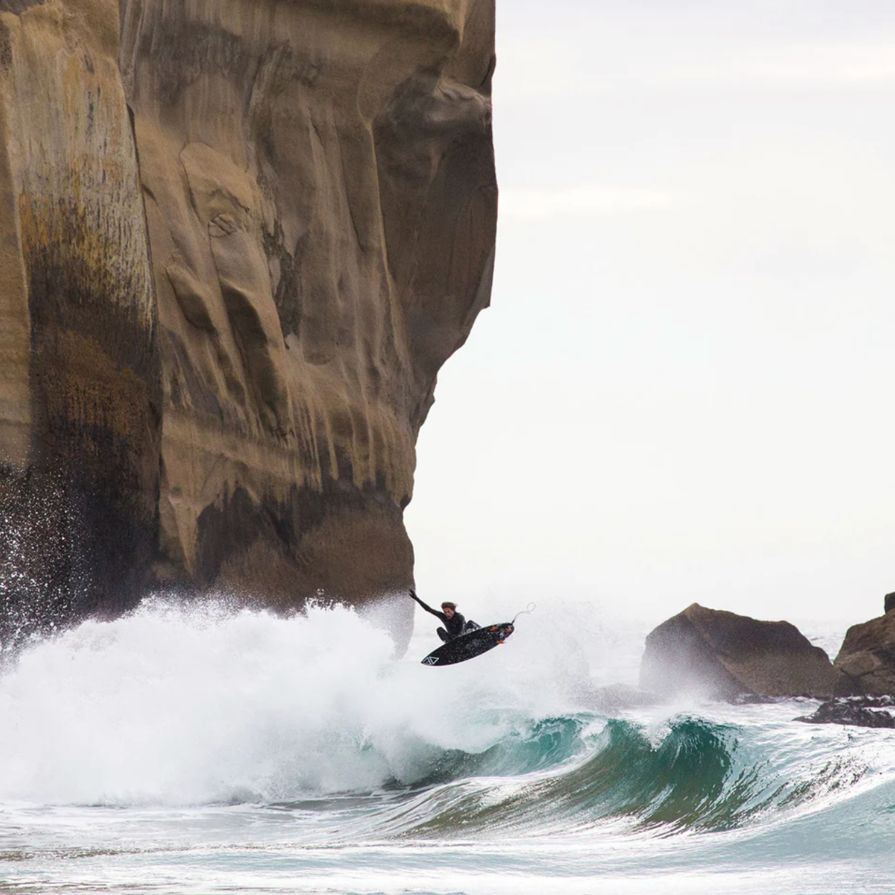 Person surfingin front of a large rock formation with a wave crashing nearby