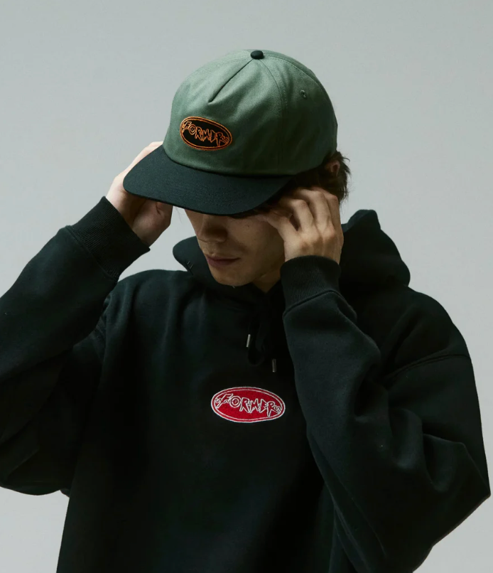 Person wearing a green cap and black hoodie with logos, against a plain background