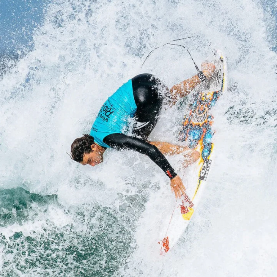 A surfer in a black and turquoise wetsuit rides a colorful surfboard through white ocean spray on a wave.