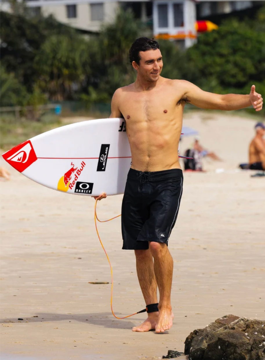 A shirtless man standing on a sandy beach holds a white surfboard branded with Red Bull, Quiksilver, and Oakley. He is wearing black swim trunks and giving a thumbs-up, with beachgoers and palm trees blurred in the background.